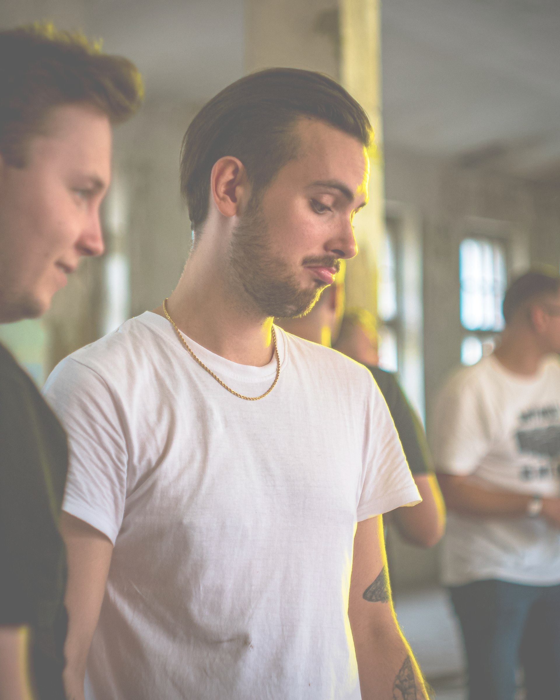 A man with a beard is wearing a white t-shirt and a gold chain around his neck.