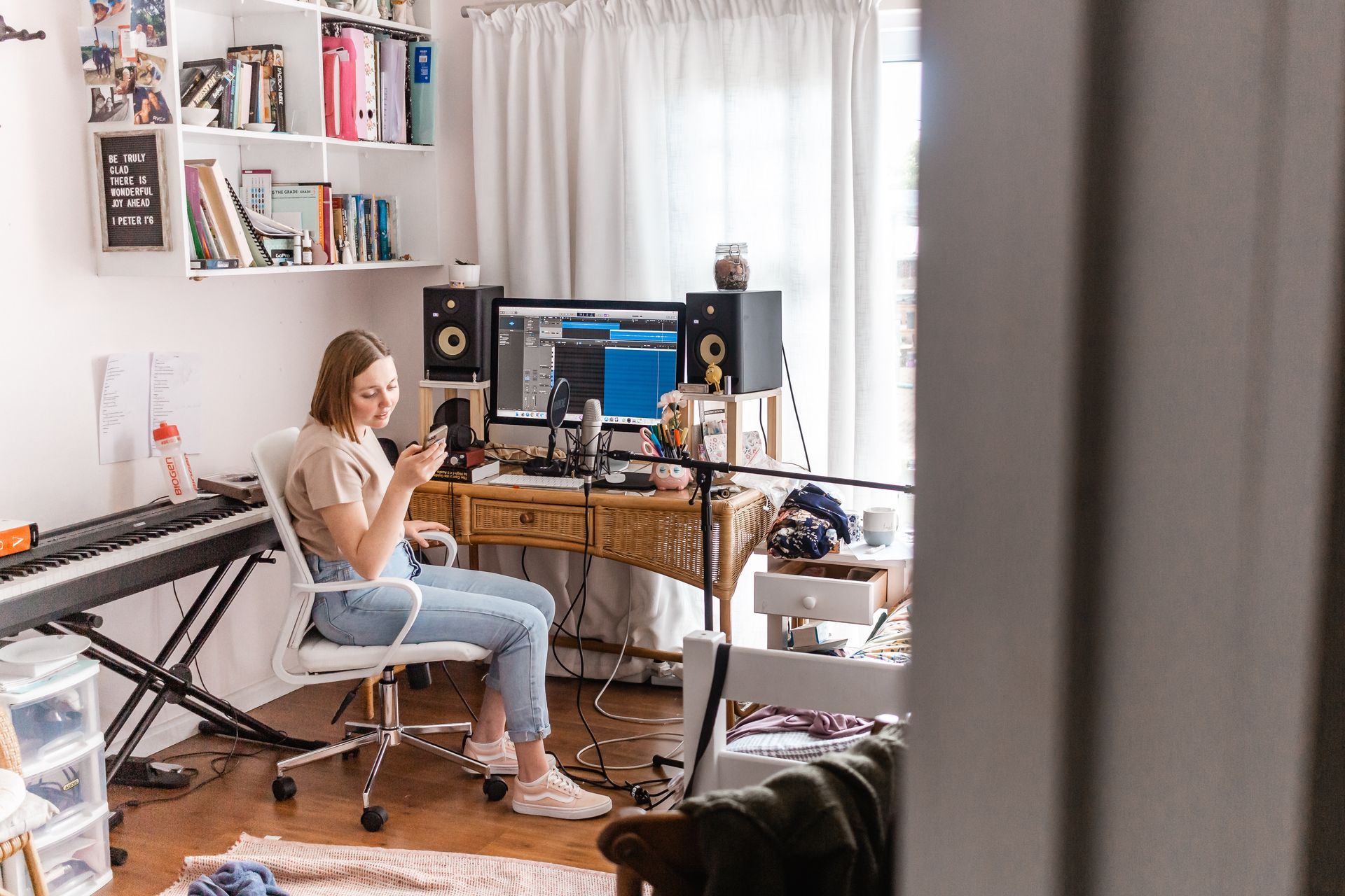 A woman is sitting at a desk in a messy room looking at her phone.