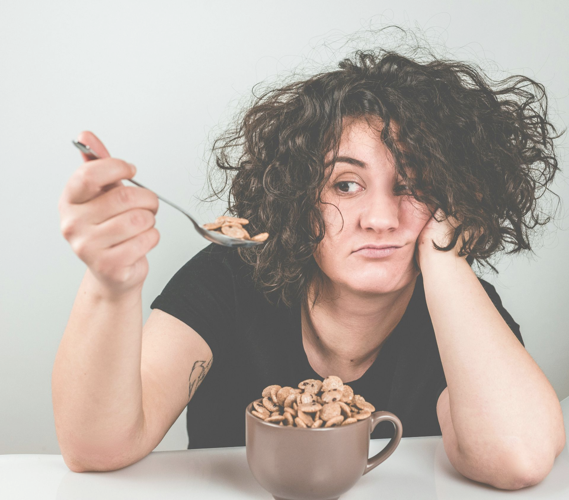 A person with curly hair holds a spoon of cereal, looking away with a bored expression while leaning on a mug of cereal.
