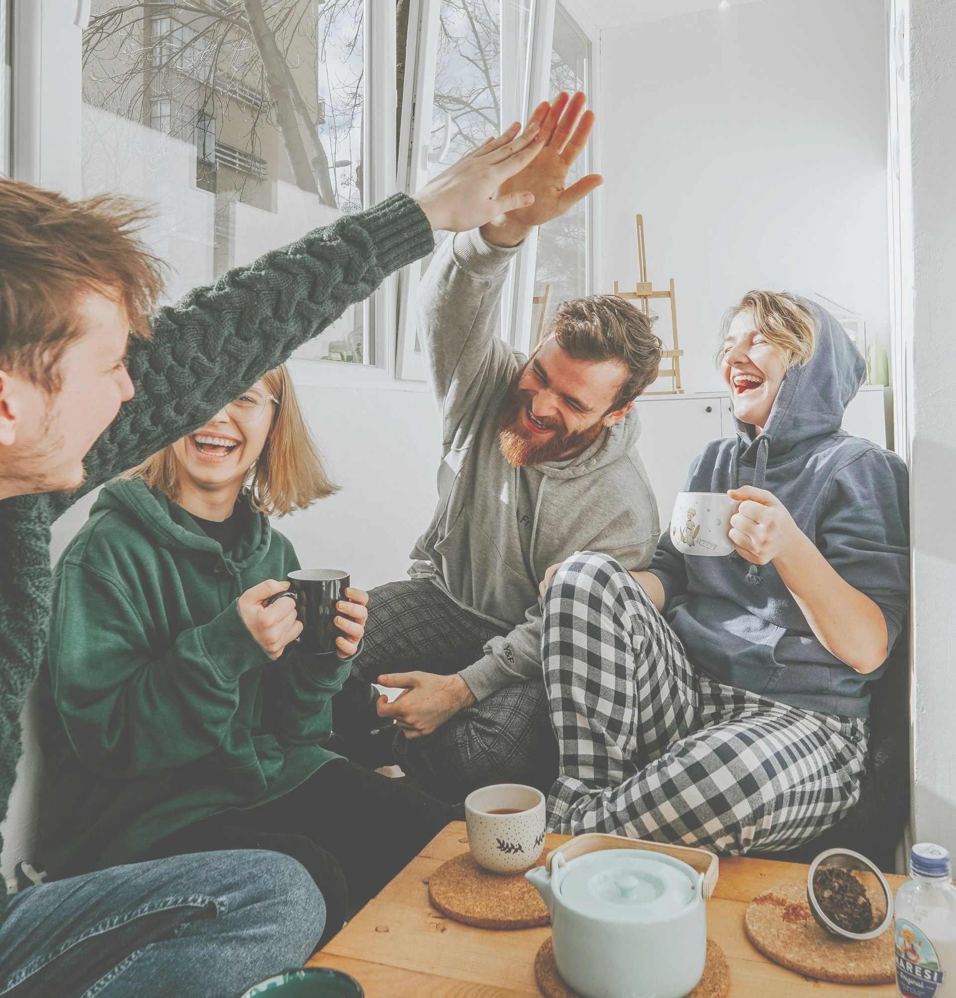 Four friends laughing, celebrating, having tea together indoors. One high-fiving.