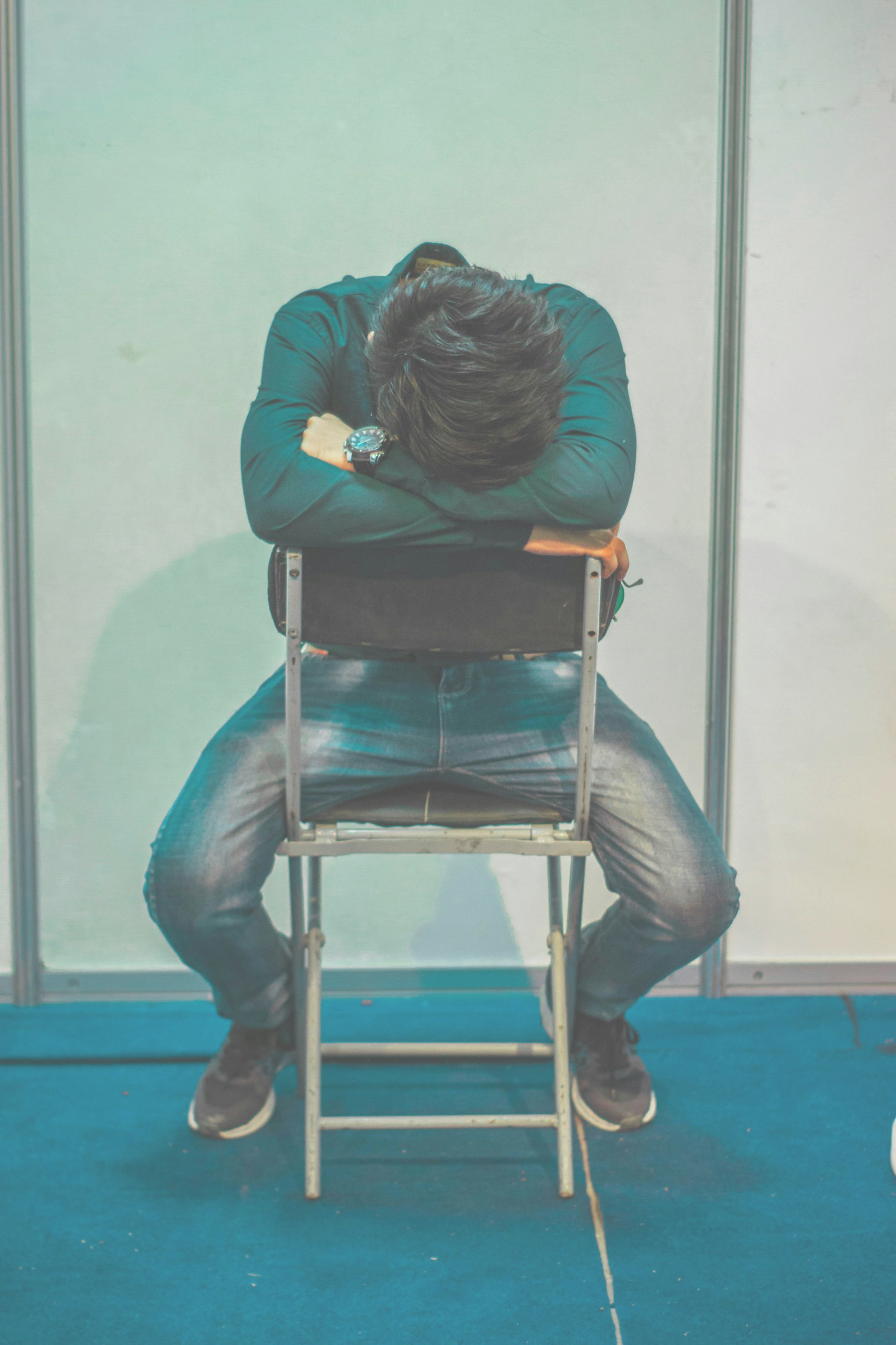 A person sits on a folding chair, head resting on crossed arms, facing away from the camera in a plain, brightly lit room.