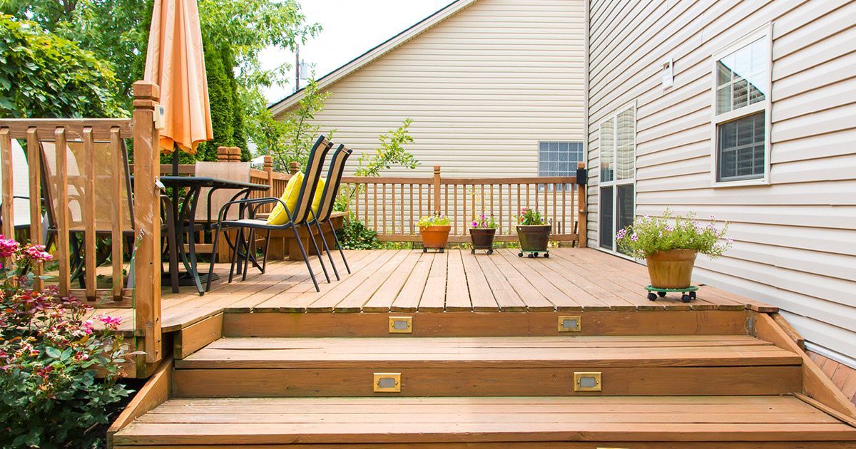 Wooden deck with steps, patio table and chairs, umbrella, and potted plants beside a house