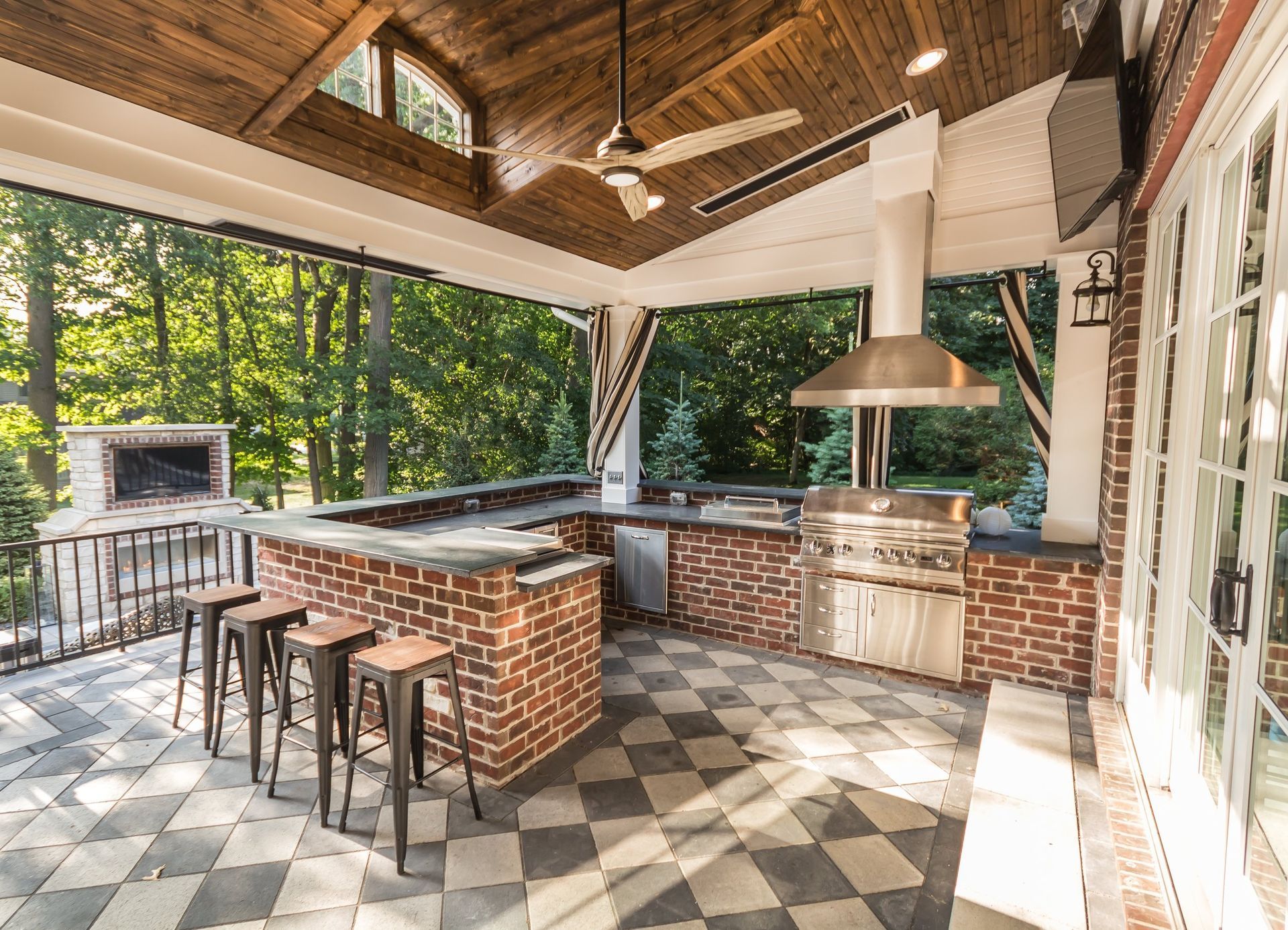 Open-air brick kitchen with bar stools, checkered floor, and large grill under a wood-beamed roof