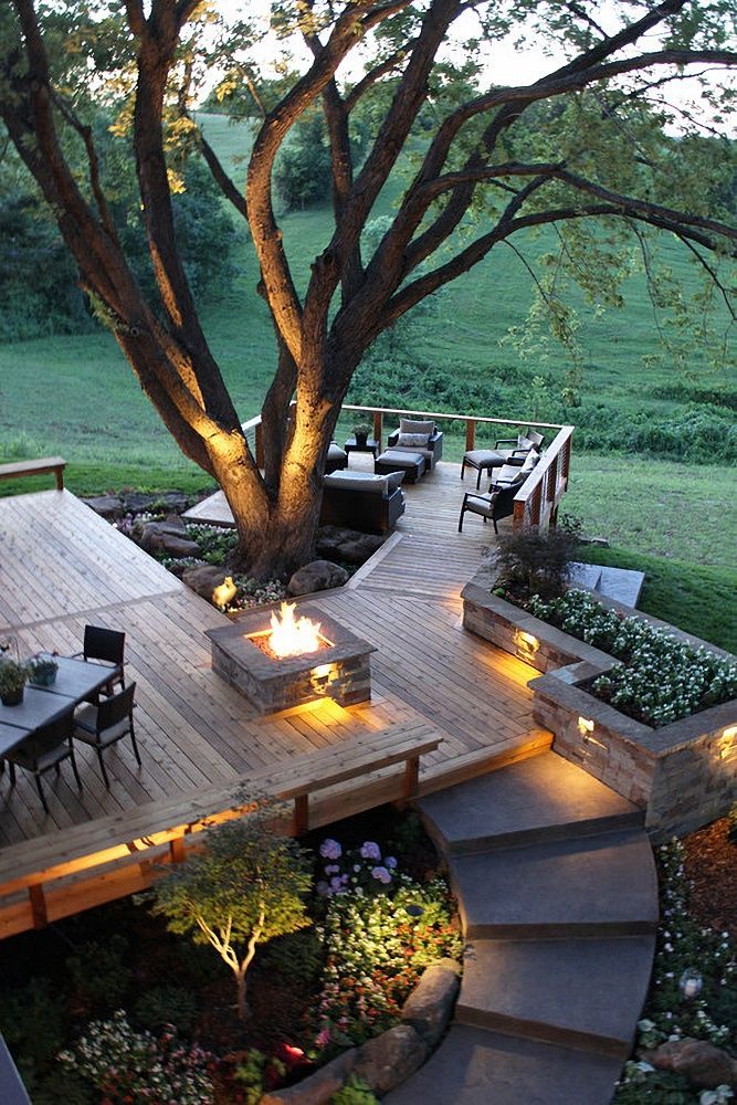Multi-level wooden deck with a large tree, fire pit, seating, and steps overlooking a green landscape