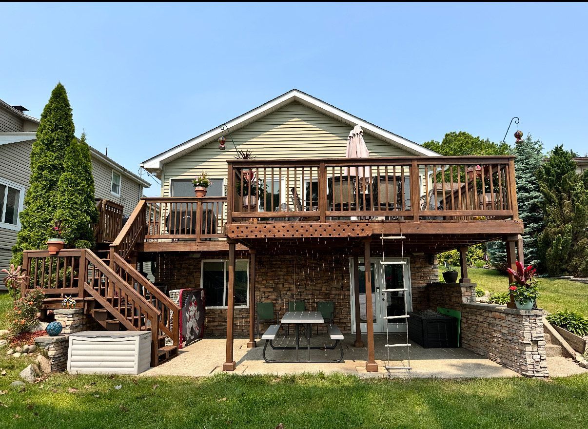 Backyard deck and patio with wooden stairs, railing, and stone supports on a house.