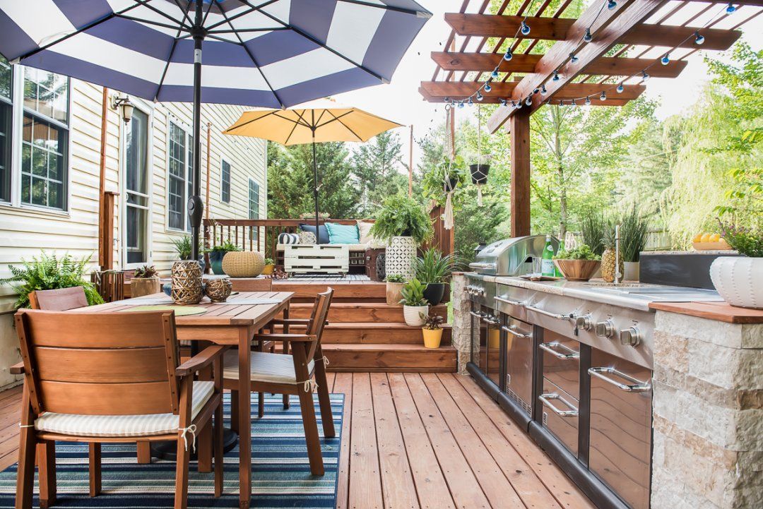 Outdoor patio with wooden dining tables, cushioned chairs, umbrellas, and a built-in grill under a pergola