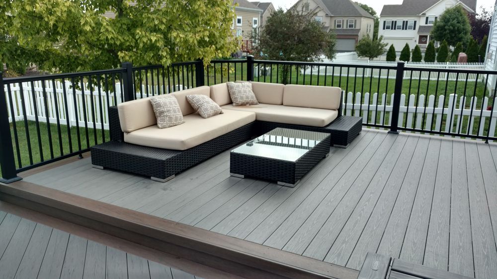 Outdoor deck with gray flooring, black railing, and beige sectional seating around a low table.