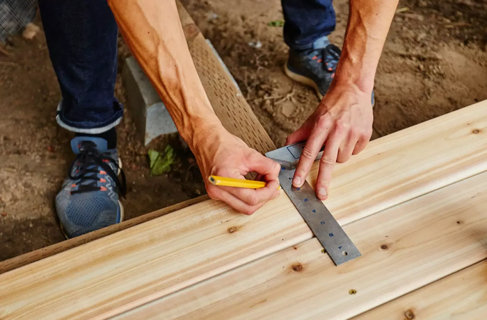 Hands measuring a wooden board with a metal square and pencil outdoors during construction