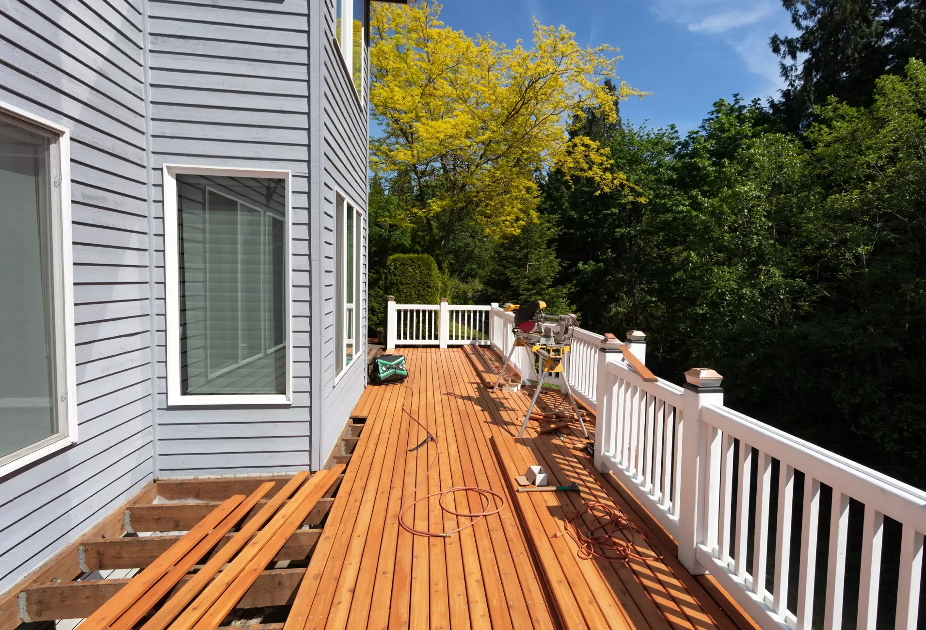 Wooden deck beside a gray house with white railing, leading toward trees under a blue sky.
