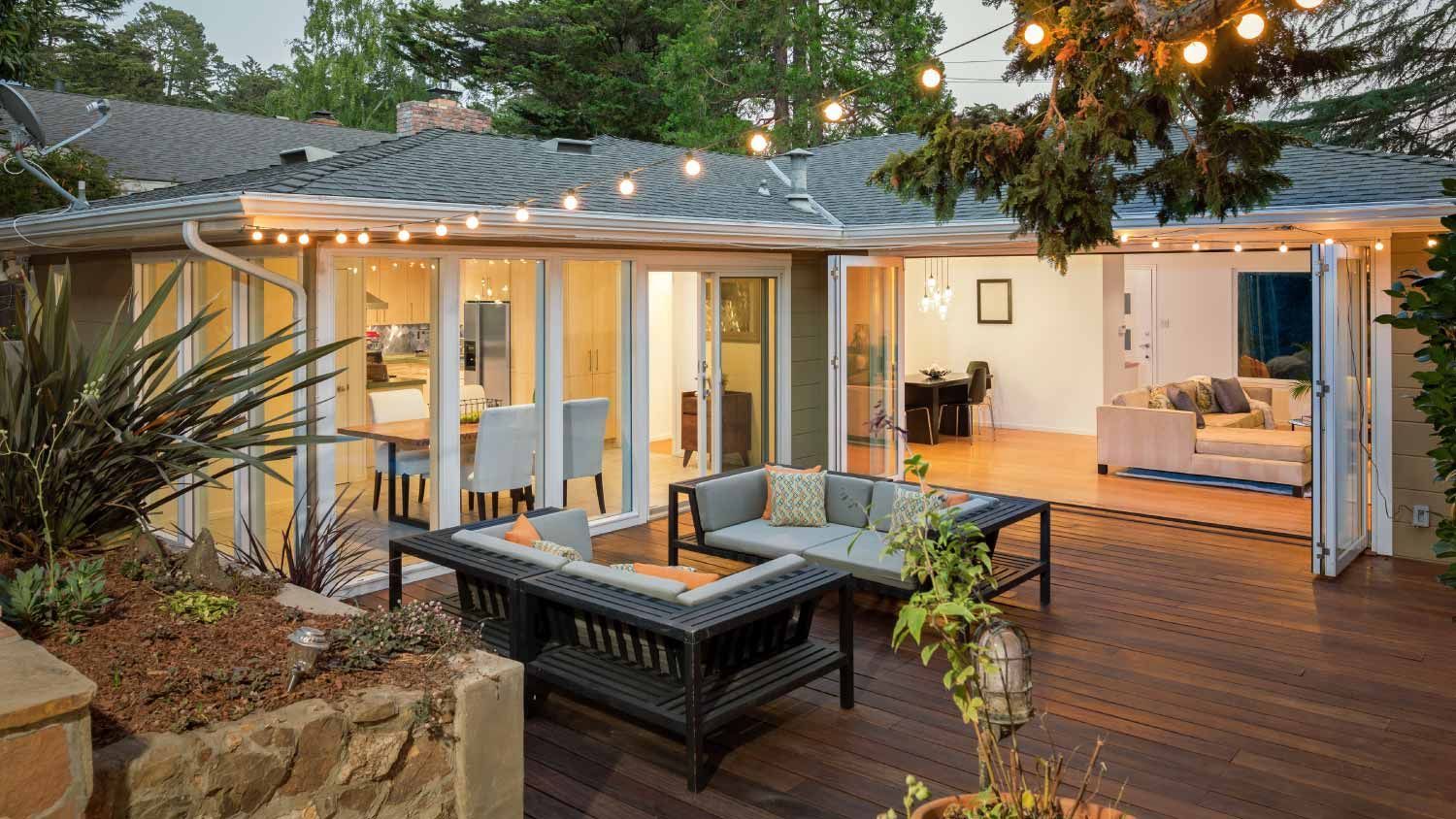 Backyard patio with wood deck, outdoor seating, string lights, and open sliding doors to a bright home interior
