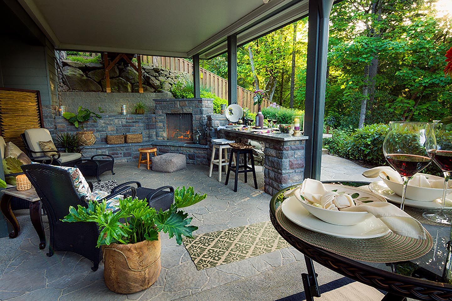 Covered patio with wicker seating, stone fireplace, and a dining table overlooking lush greenery