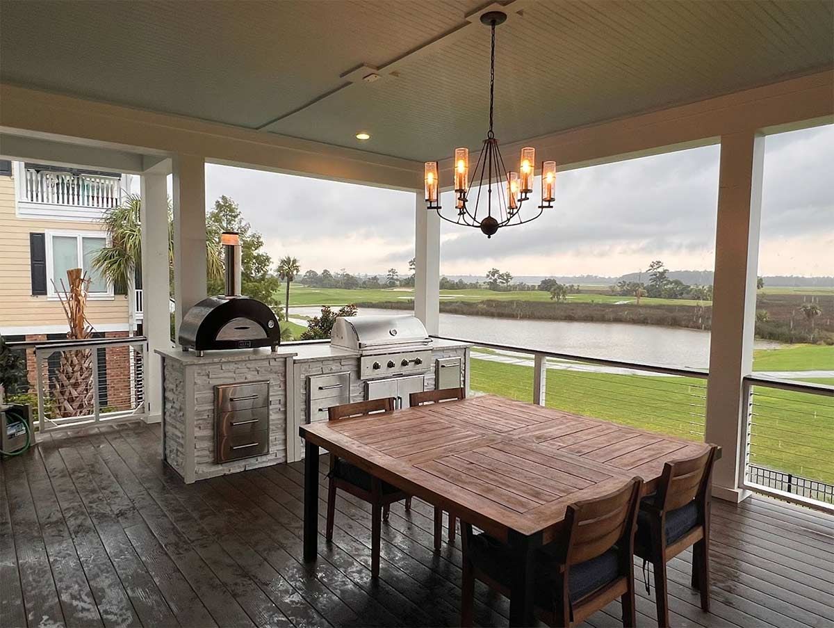 Covered patio dining area with table, chairs, grill, and chandelier overlooking a pond and green lawn