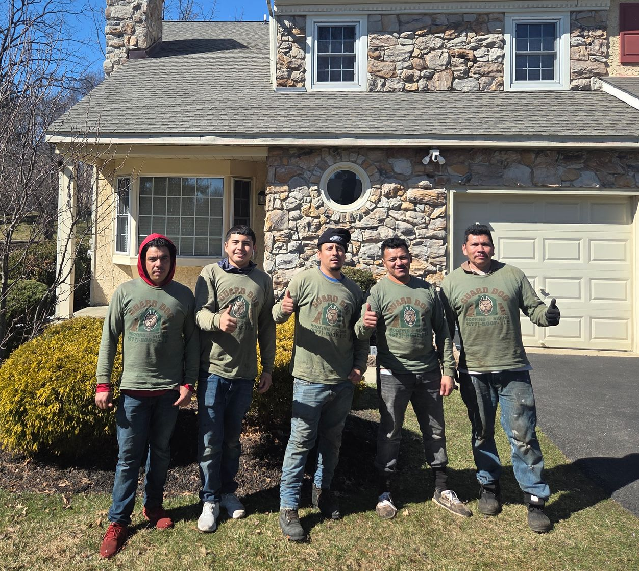 Five people standing in front of a stone house on a sunny day, wearing matching green shirts.