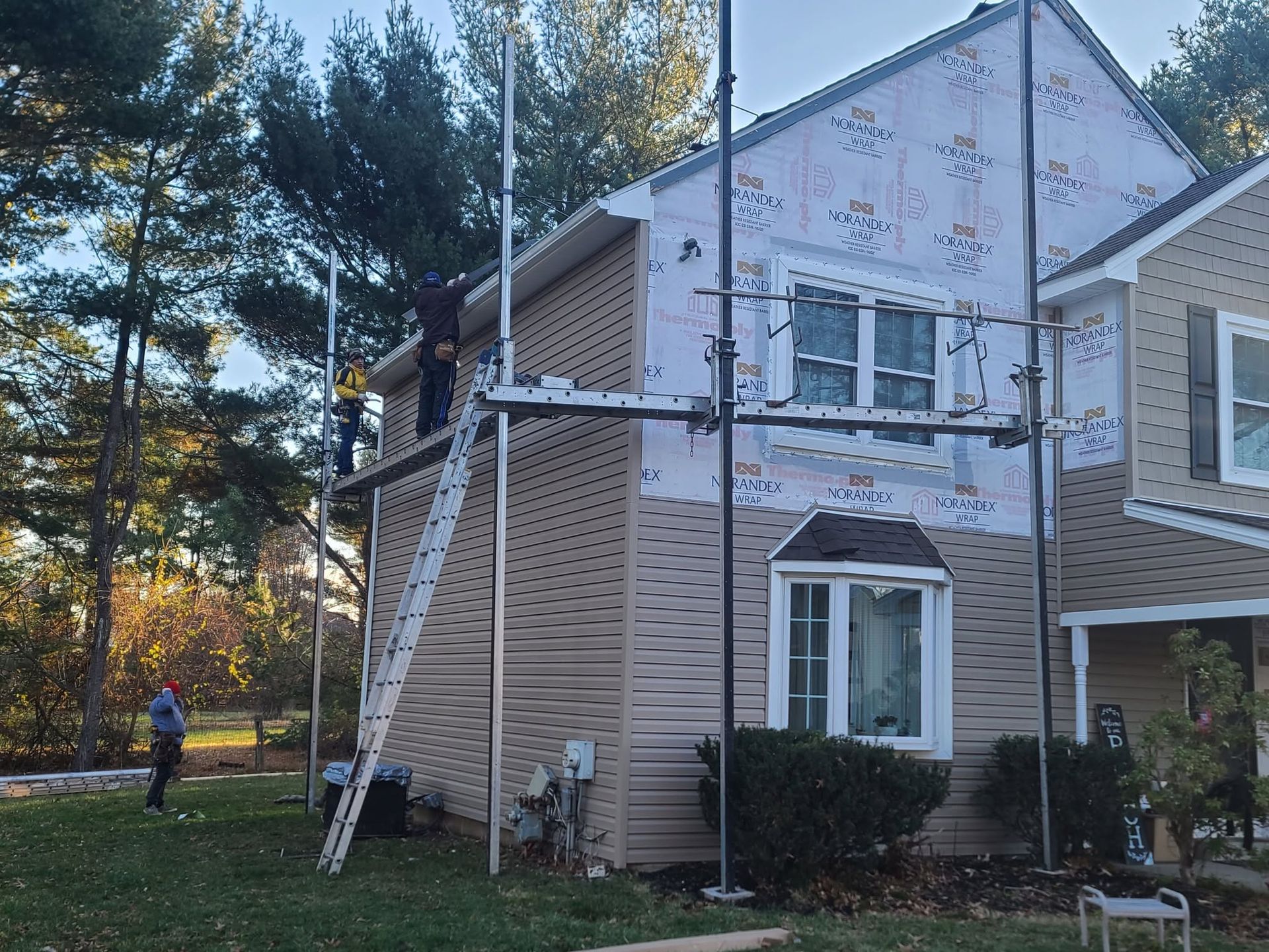 House exterior under repair with scaffolding and a worker nearby, surrounded by trees in a yard