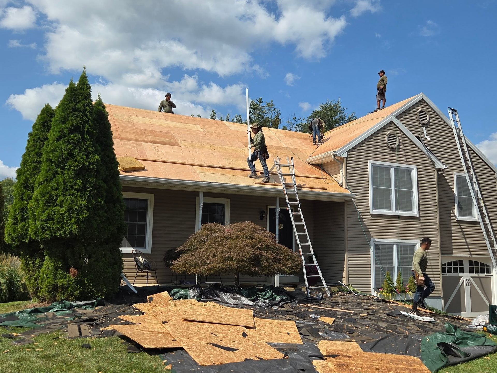 Workers reroofing a gray house with tan shingles under a blue sky