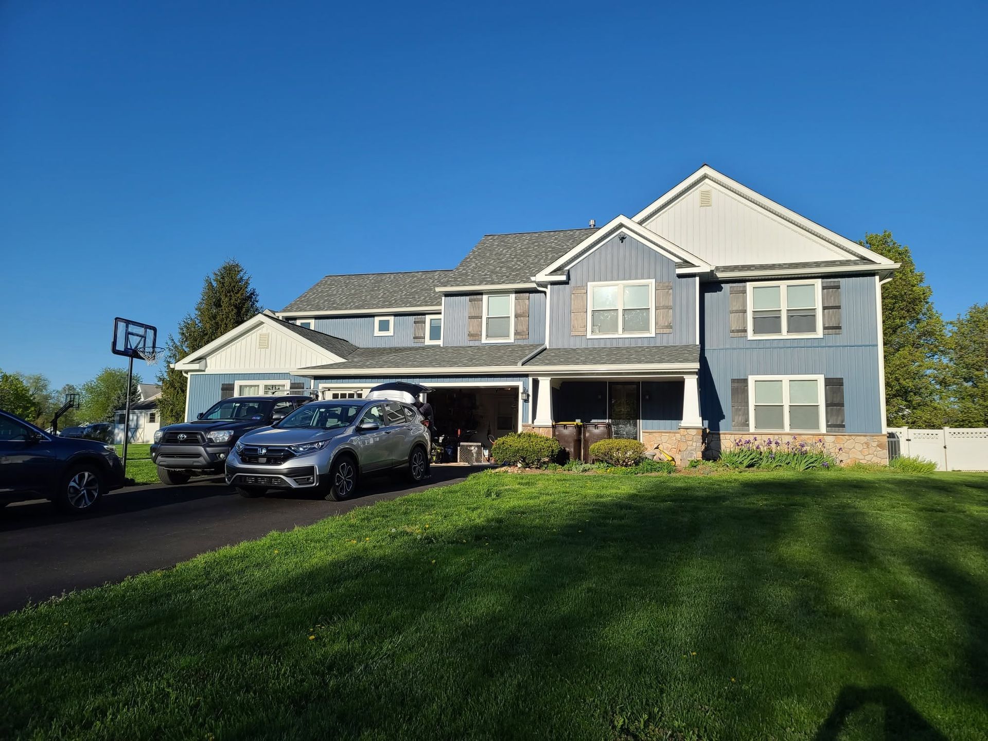 Suburban two-story gray house with a driveway, parked car, and a green lawn under a blue sky