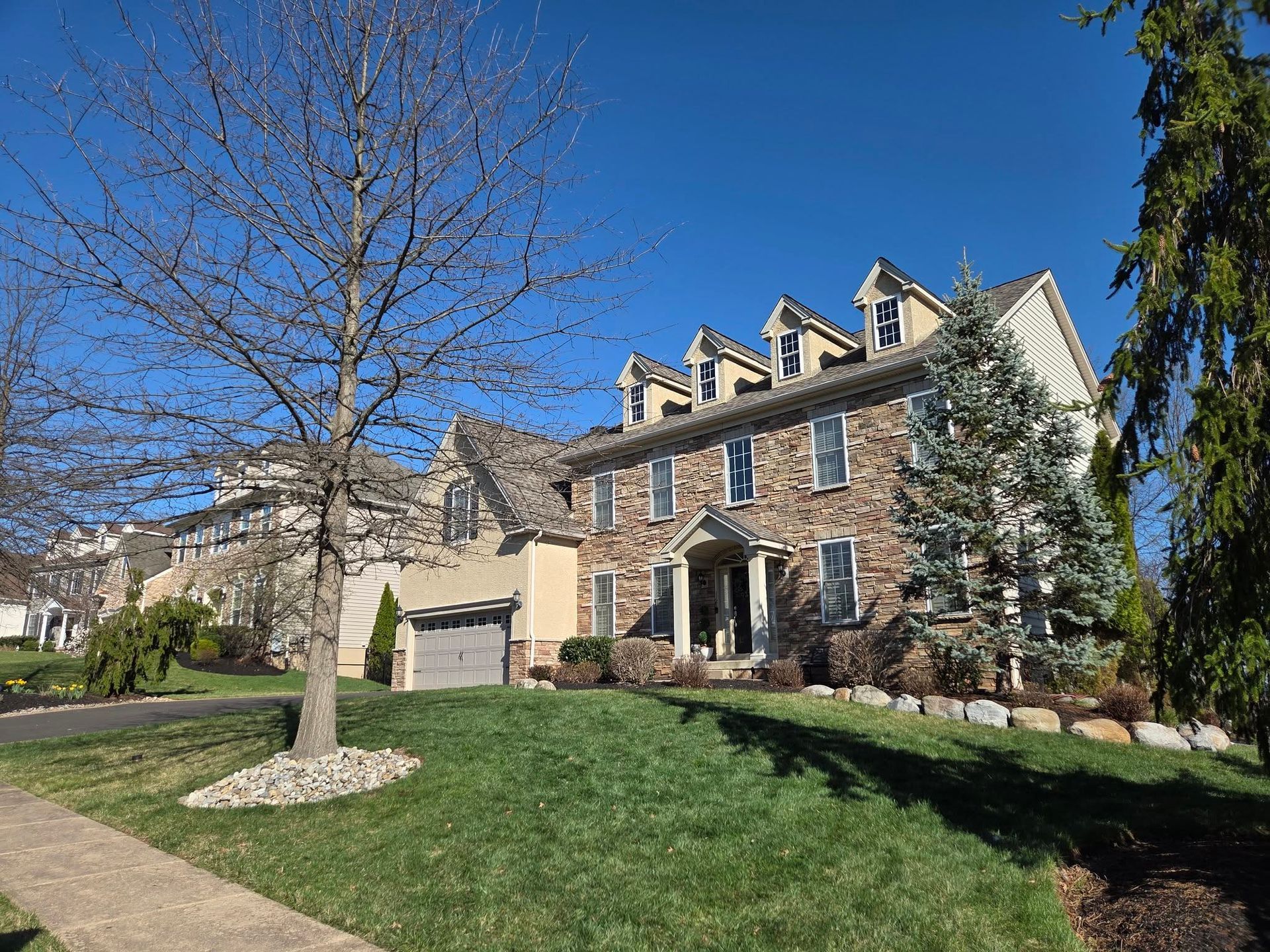 Brick suburban house with dormer windows, front lawn, and leafless tree under a clear blue sky