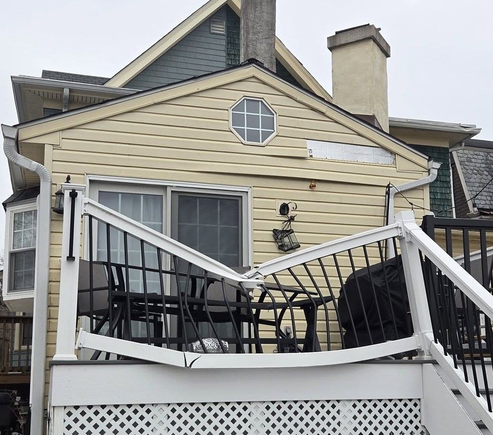 Damaged deck railing on a beige house after storm or impact, with bent white posts and black patio furniture