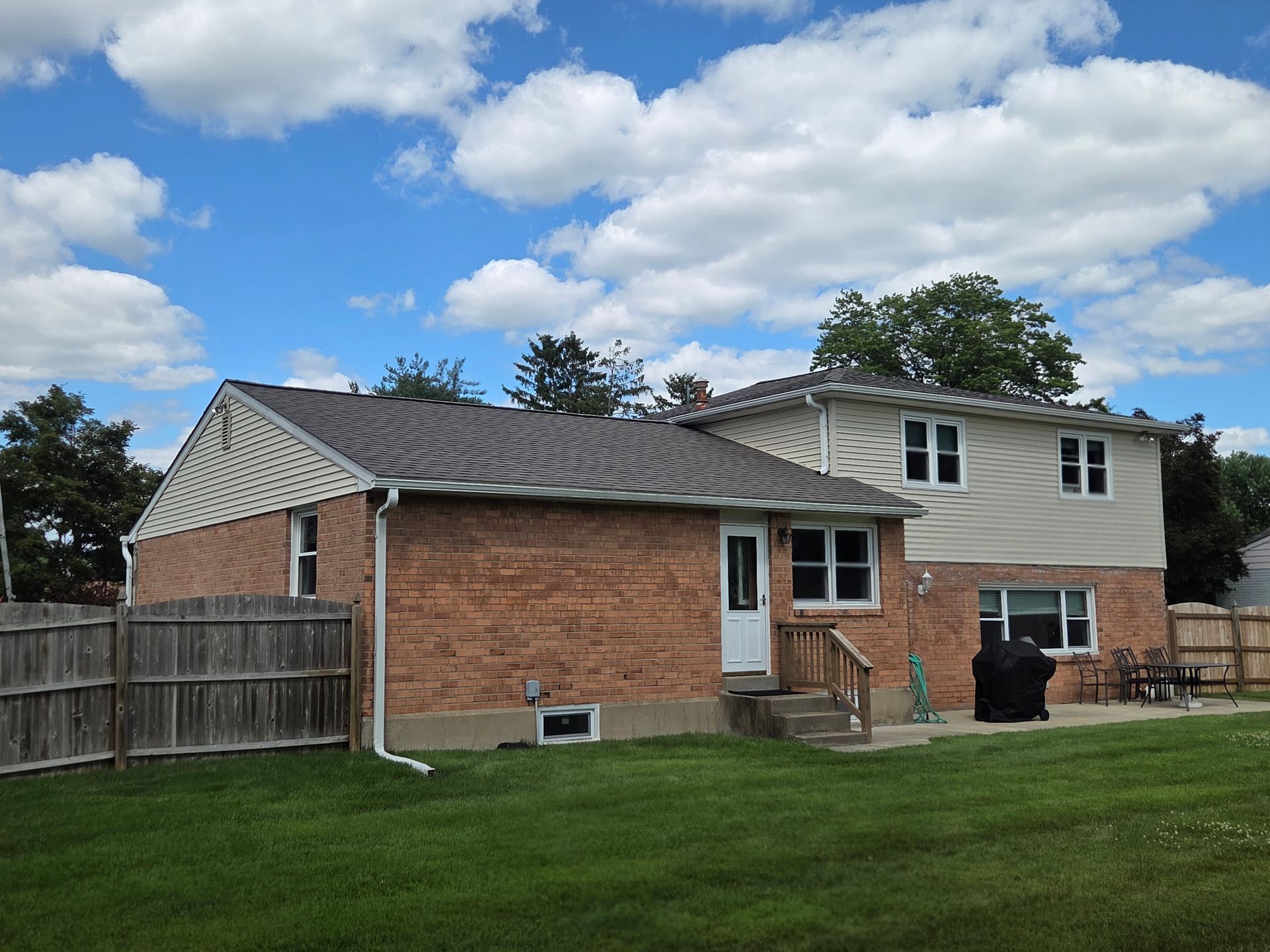 Backyard with a brick-and-siding house, green lawn, wooden fence, and partly cloudy sky