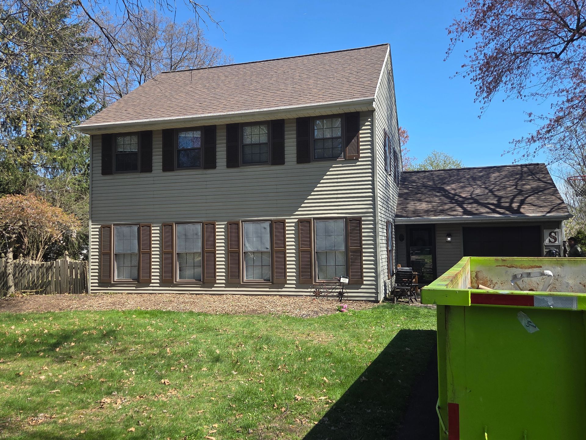 Two-story gray house with brown shutters and a green dumpster in the foreground