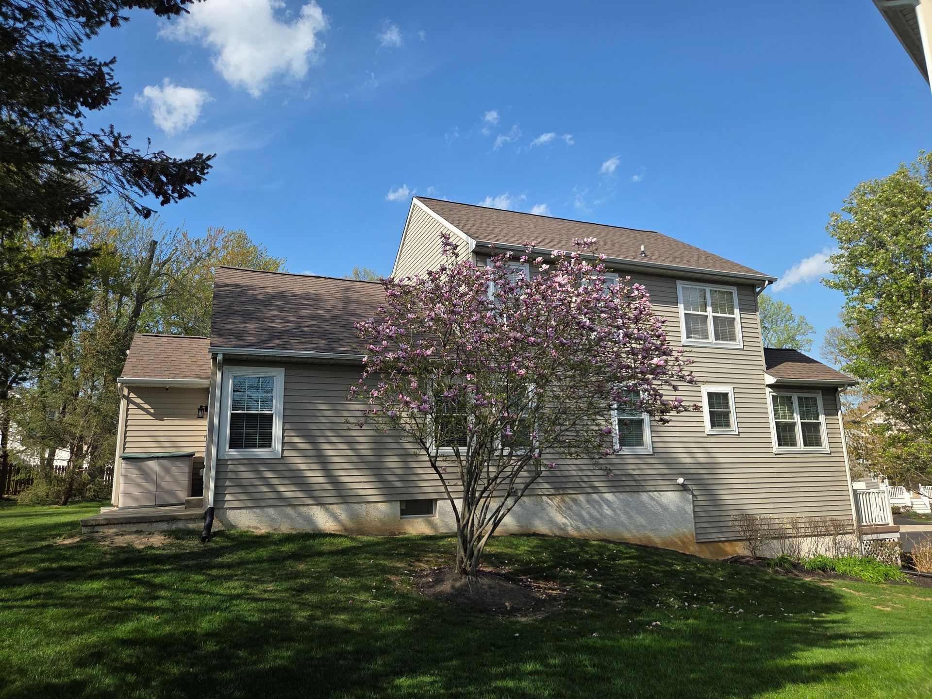 Two-story beige house with a flowering tree in the front yard under a blue sky
