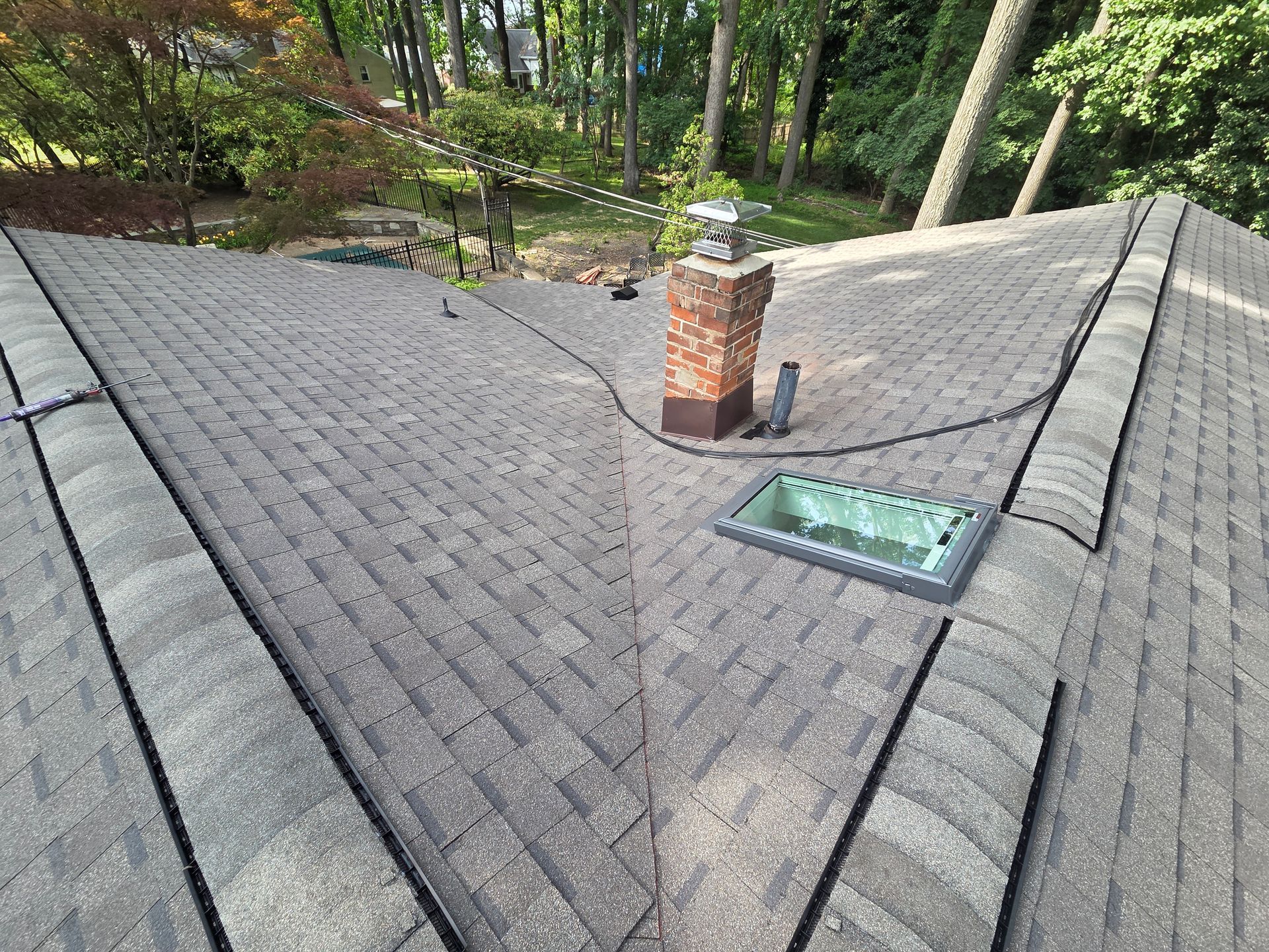 Shingled roof with a brick chimney and skylight, surrounded by trees.