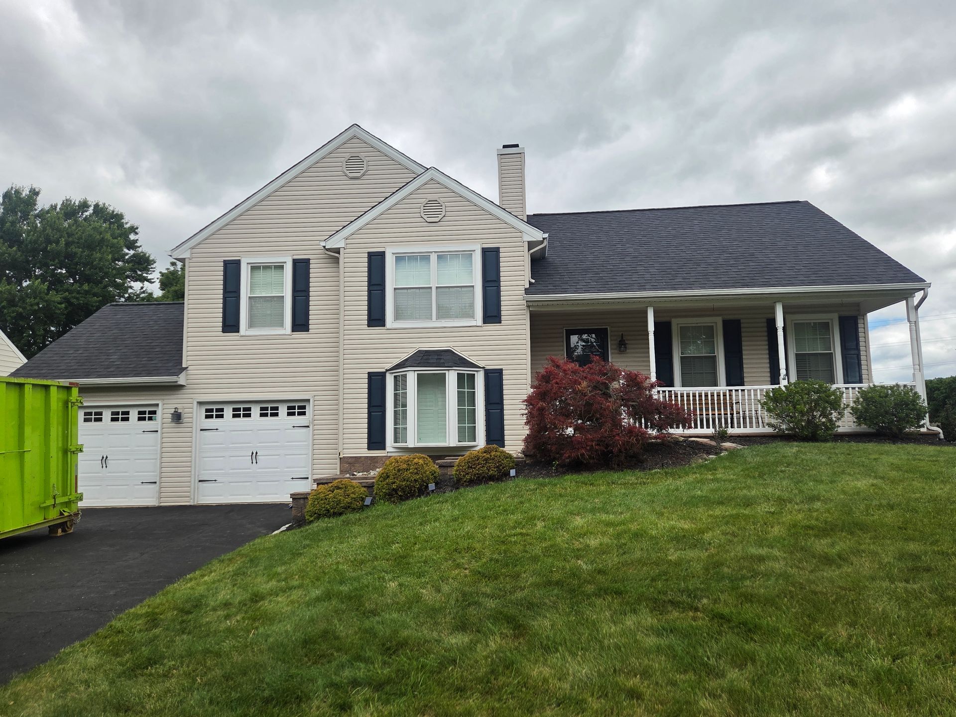 Two-story beige house with white trim, green shutters, and a garage on a sloped lawn under cloudy sky