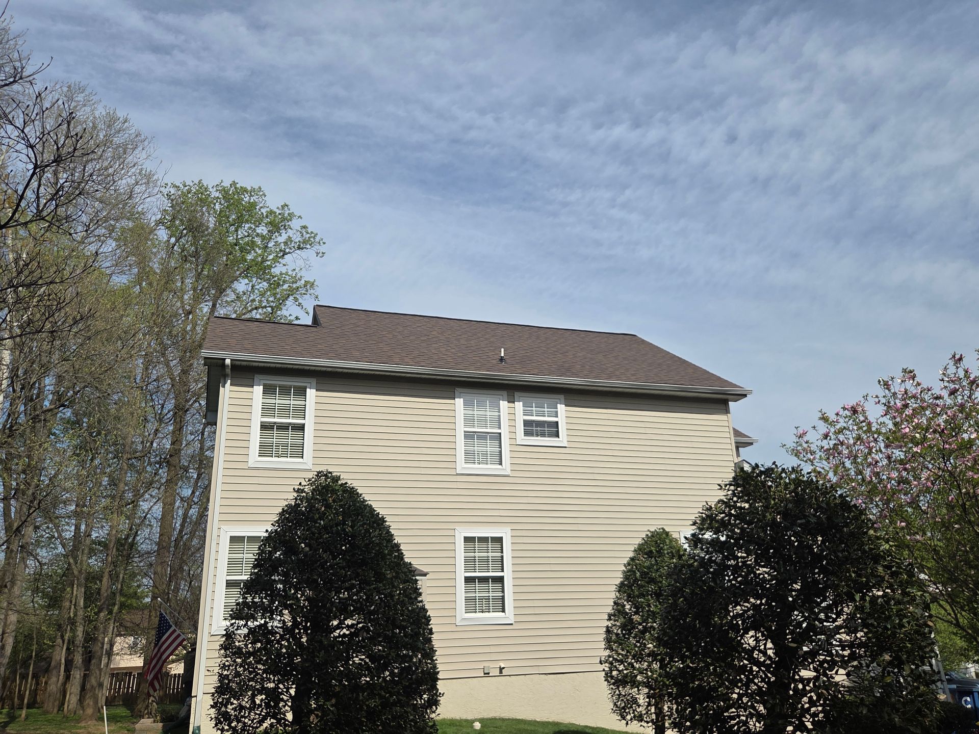 Two-story beige house with a dark roof, framed by trees under a cloudy sky