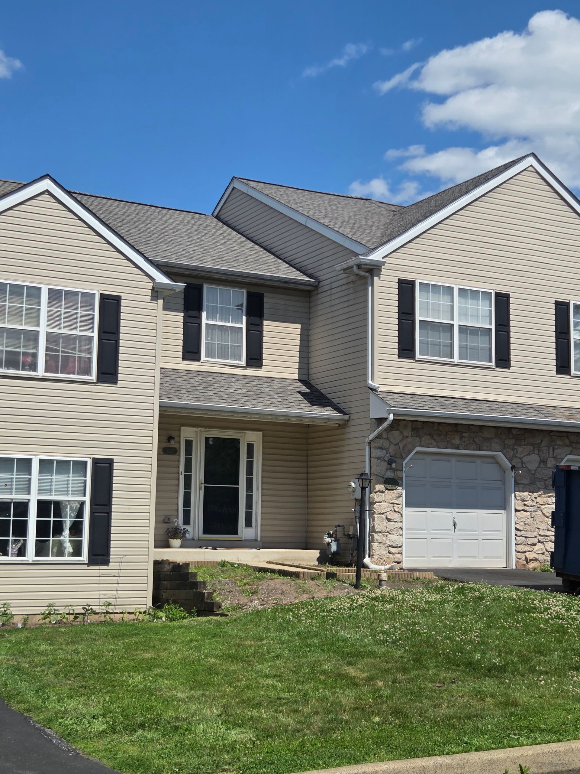 Suburban beige two-story house with black shutters, white garage, and green lawn under a blue sky