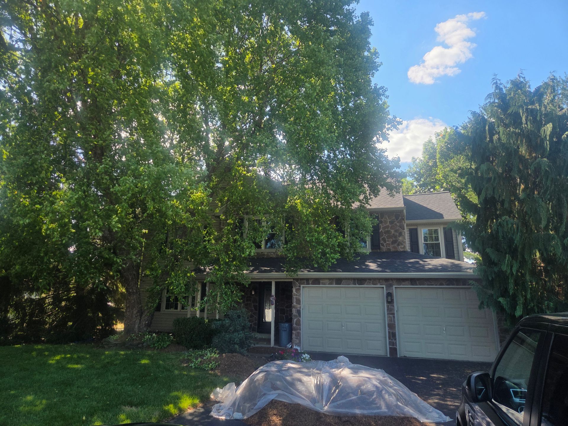 Tree-shaded suburban house with a two-car garage and a tarp-covered object in the driveway