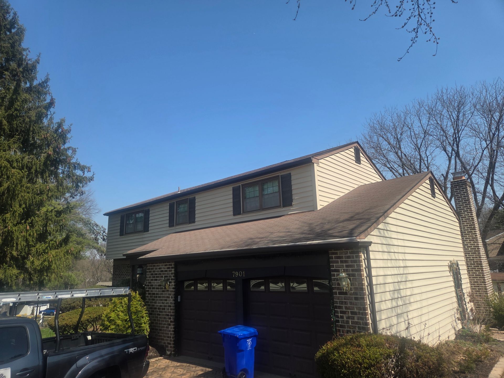 Two-story tan house with attached garage, blue trash bin, and trees under a clear sky.