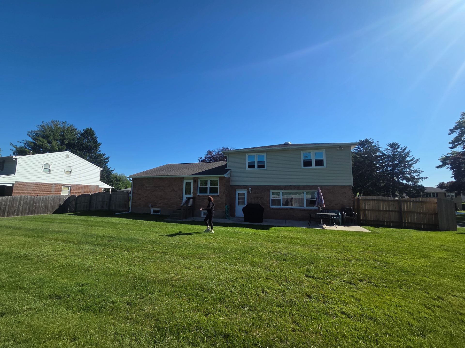 Suburban backyard with two-story brick house, wooden fence, and a person standing on a sunny lawn