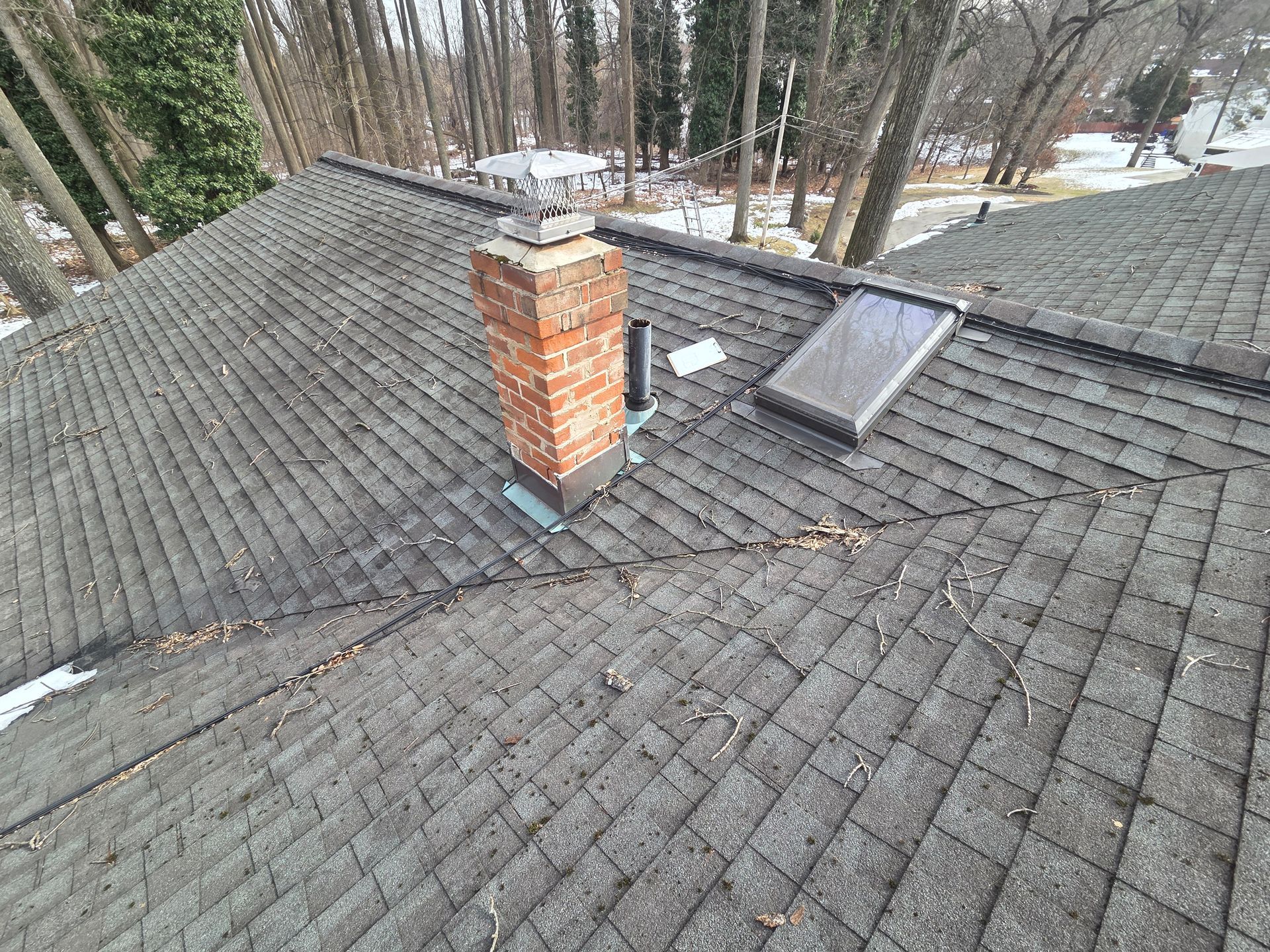 Gray shingled roof with a red brick chimney and skylight amid trees and snow