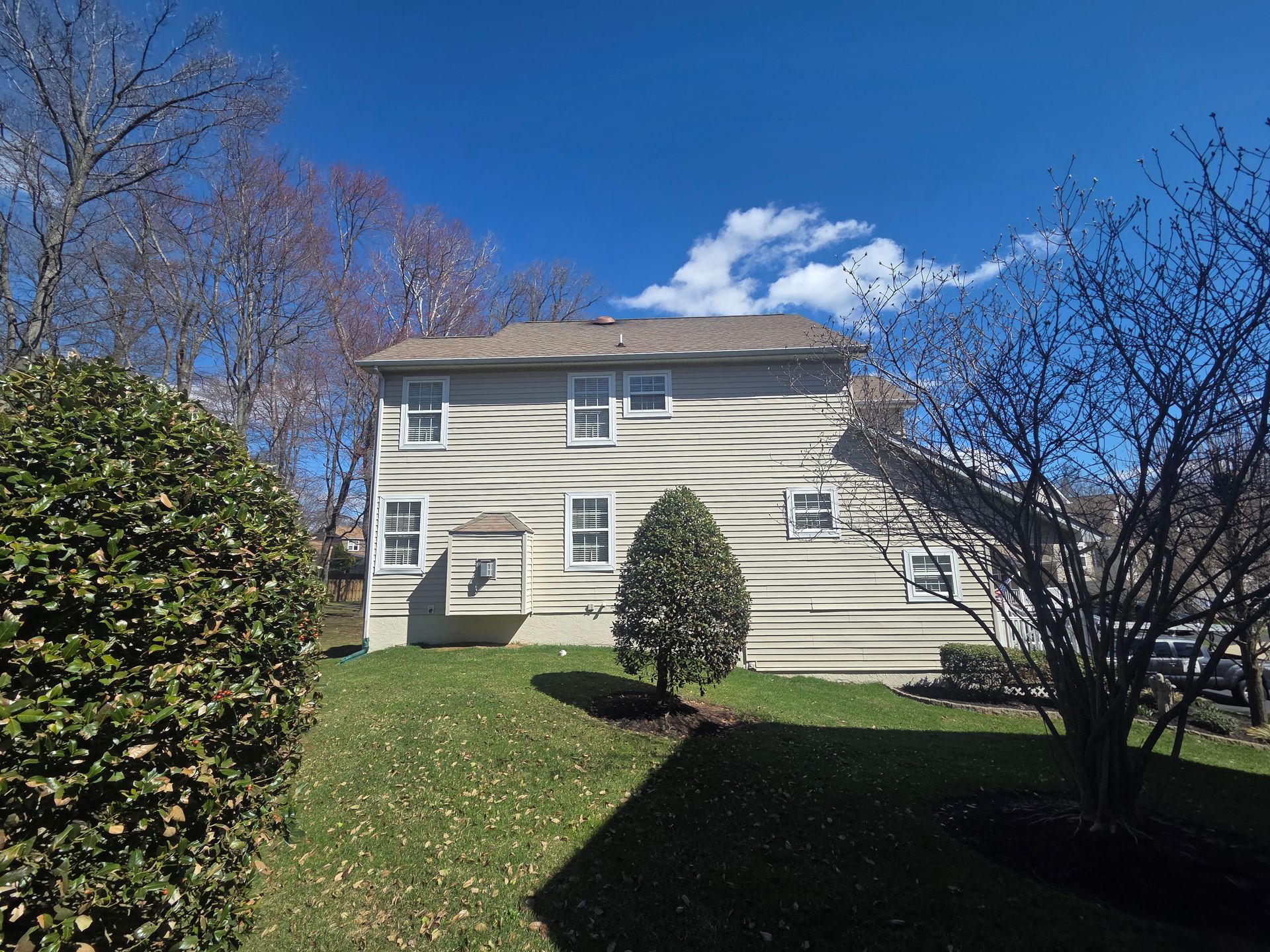 Two-story beige house with lawn, trees, and blue sky on a sunny day