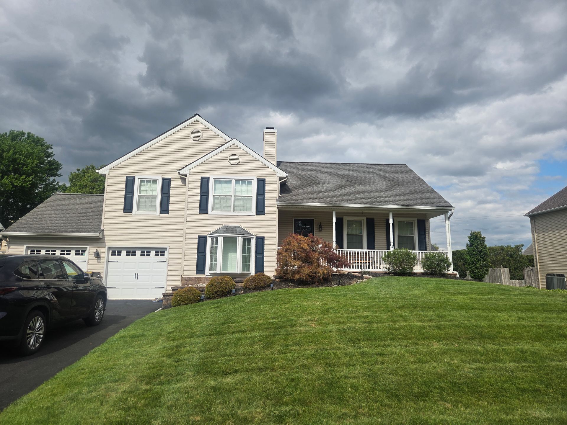 Suburban two-story beige house with a garage, porch, and green lawn under cloudy skies