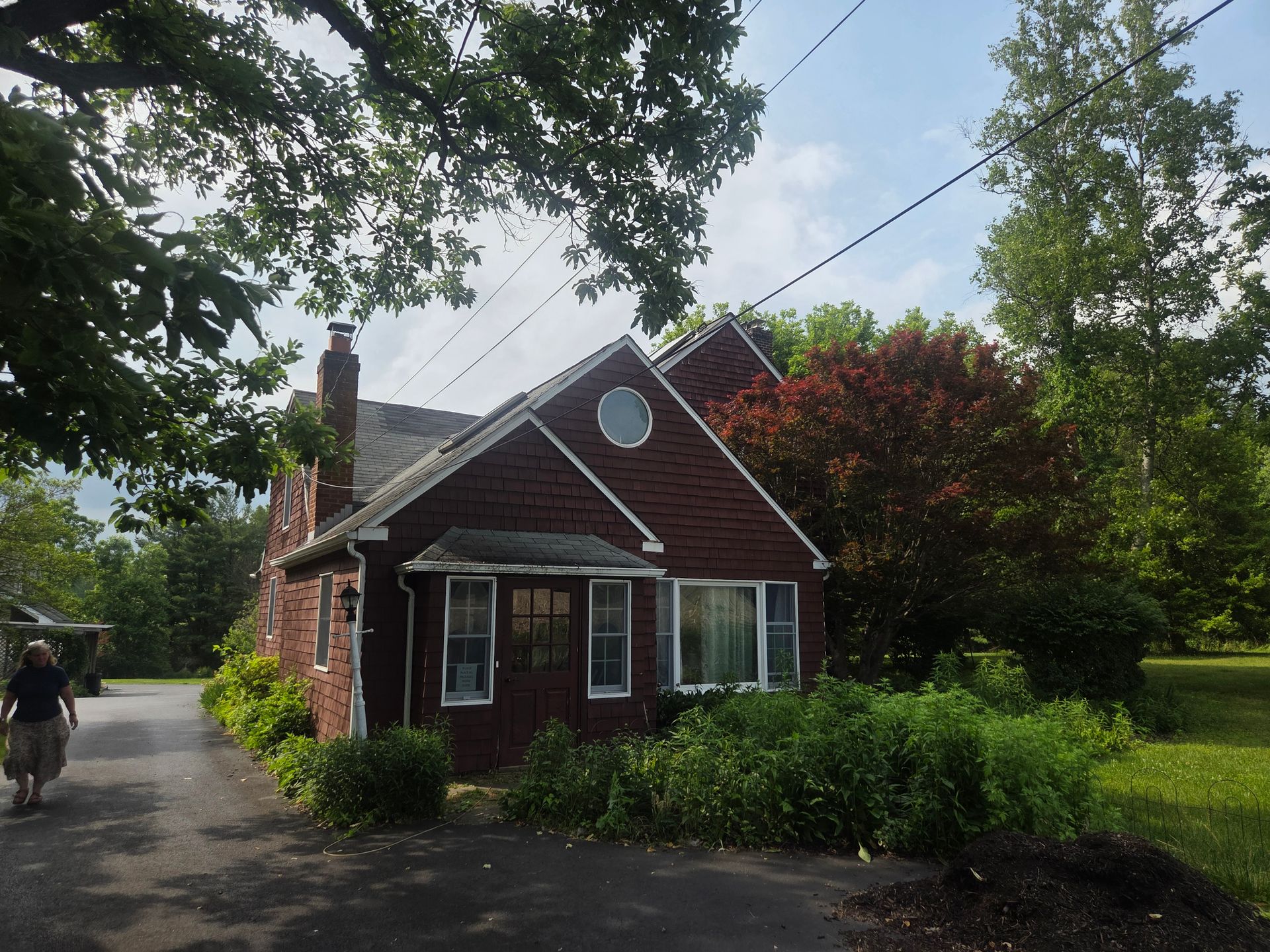 Small brick house with a steep roof and round window, set among trees and shrubs beside a driveway.