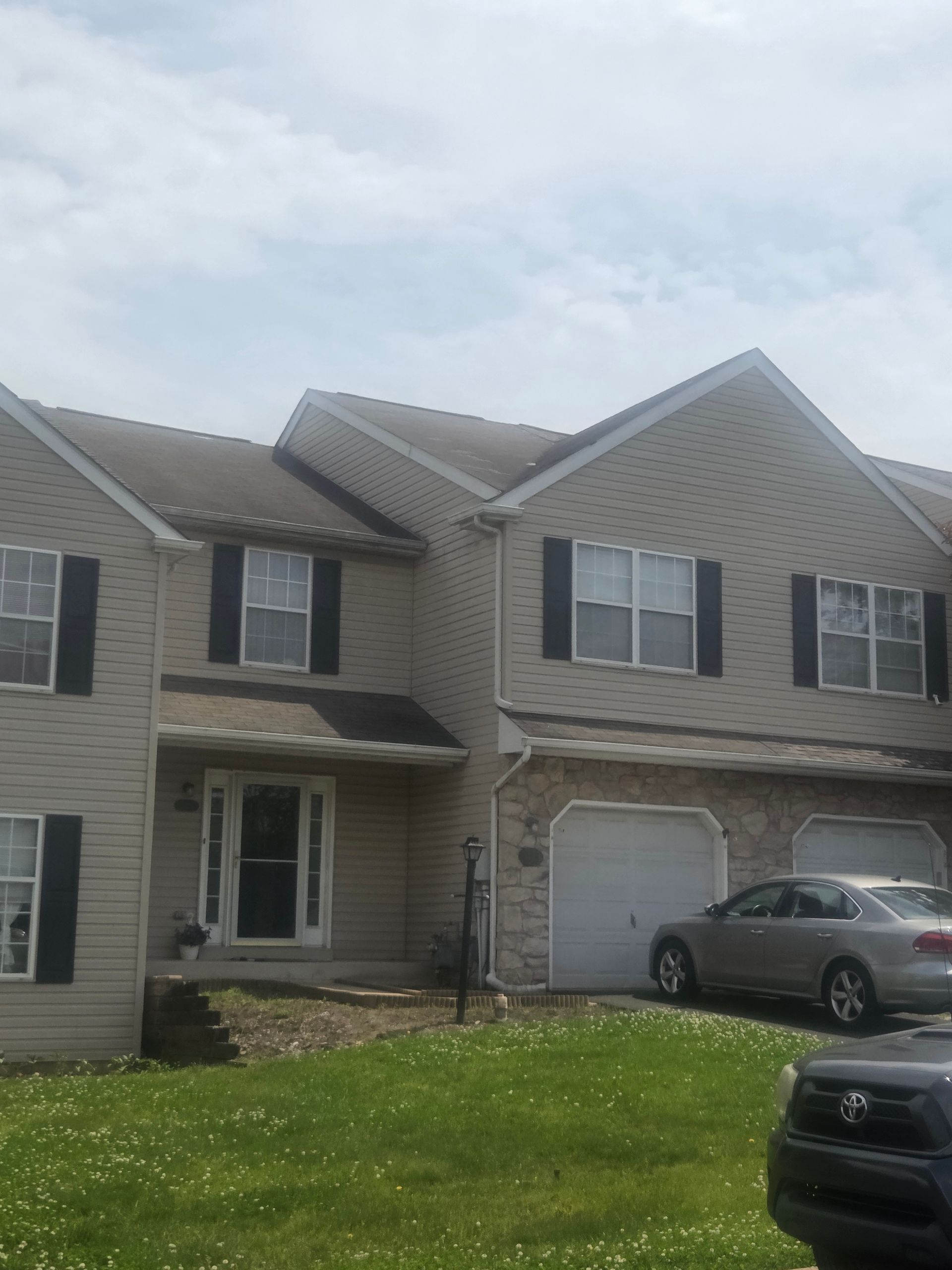 Suburban two-story beige house with attached garage, front porch, and cars in the driveway under cloudy skies
