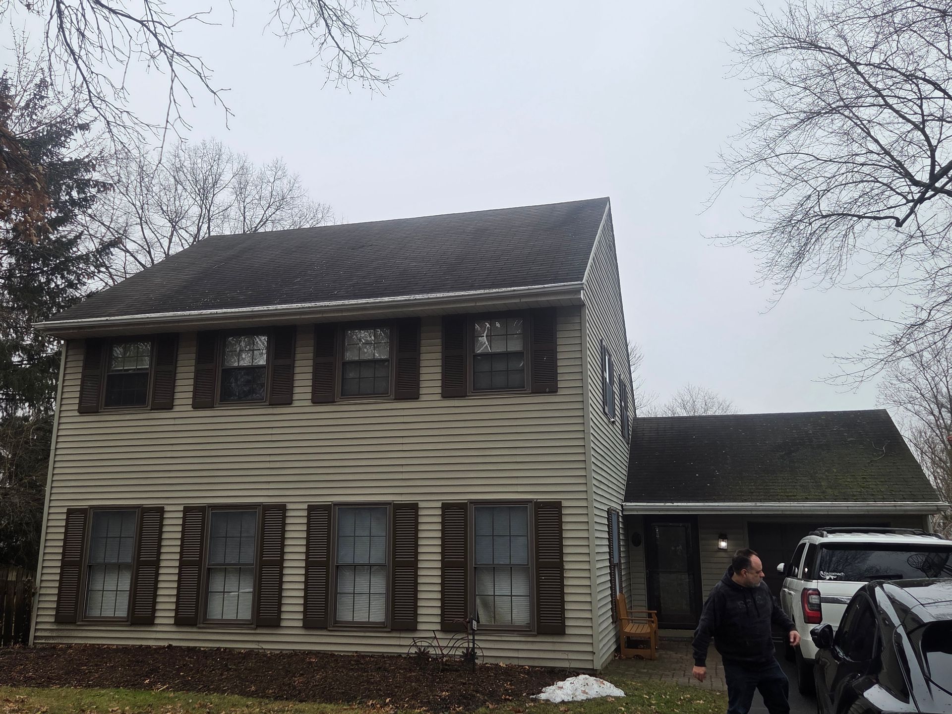 Two-story house with striped siding and a carport; a person stands near a parked vehicle on a cloudy day.