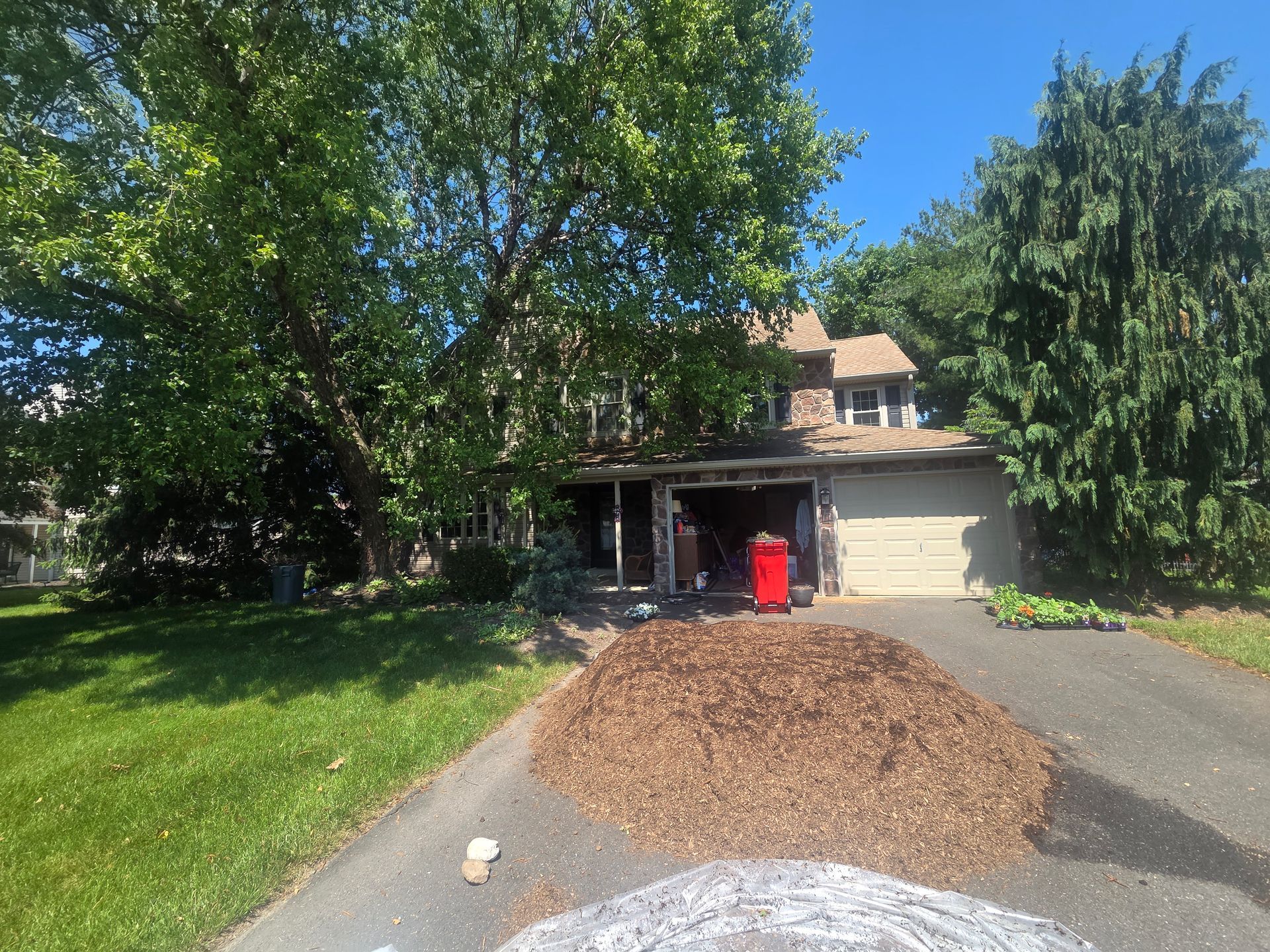 Driveway piled with mulch beside a house, with a red bin and leafy trees on a sunny day