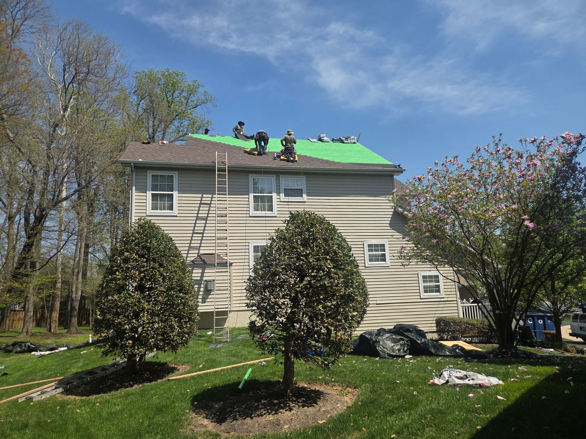 House with workers on a green roof, surrounded by trees and lawn under a blue sky