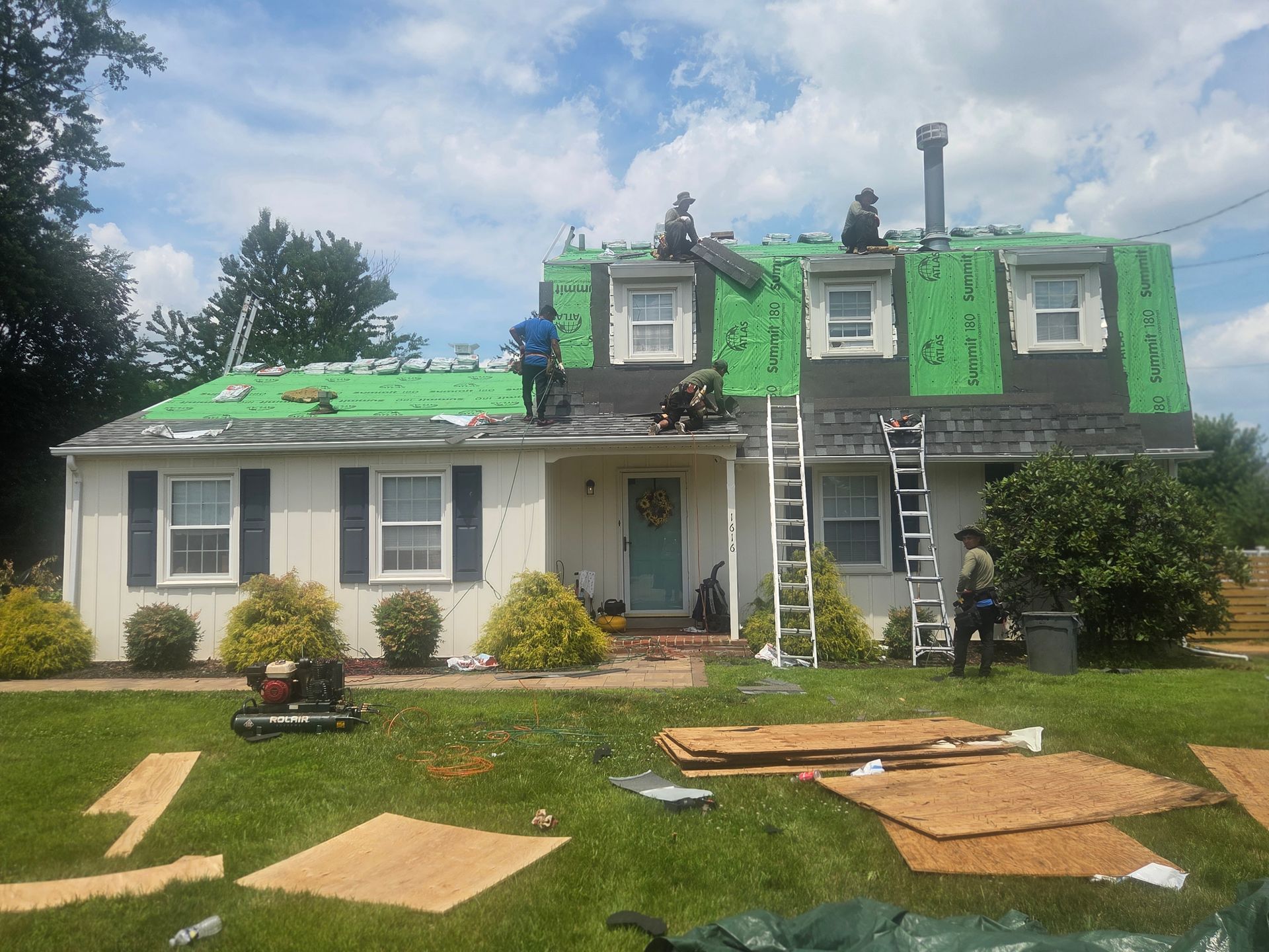 House roof being repaired with green tarp and ladders; shingles and materials scattered on the front lawn.