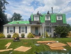 House roof under repair with workers on ladders and green protective coverings, debris and boards on the lawn