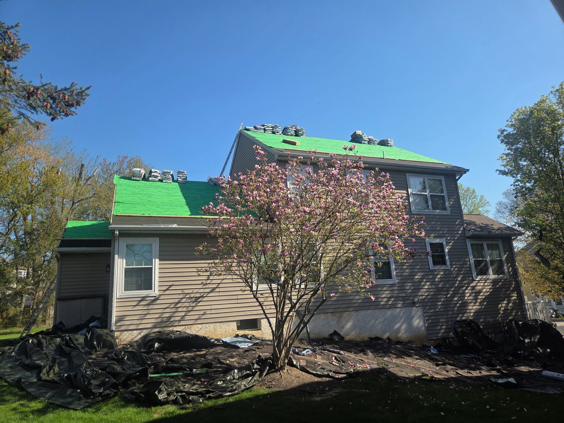 House with green roof tarp amid storm damage and fallen debris under a clear blue sky