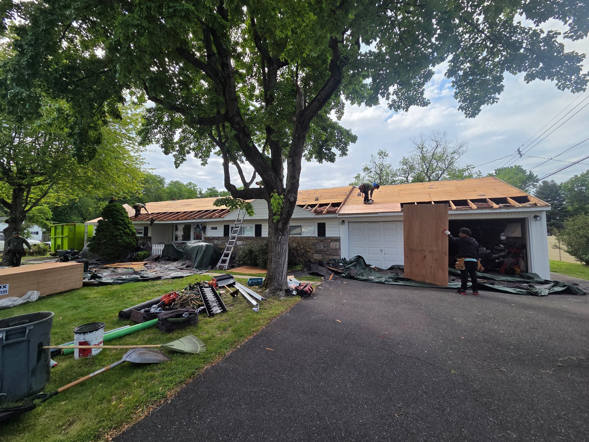 Driveway with a large tree, scattered tools, and a house with a partially repaired roof and garage.