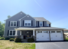 Gray two-story suburban house with white trim, front porch, and a two-car garage under a clear blue sky