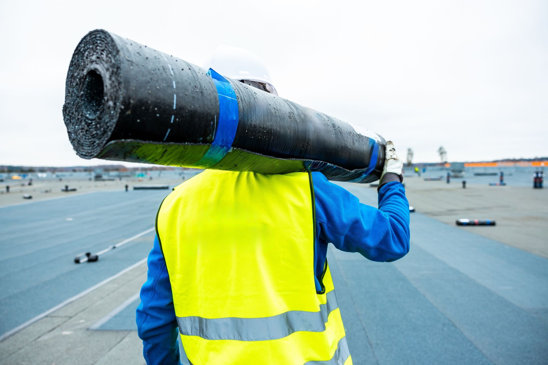 Worker in a neon yellow vest carrying a rolled black roofing membrane on a rooftop