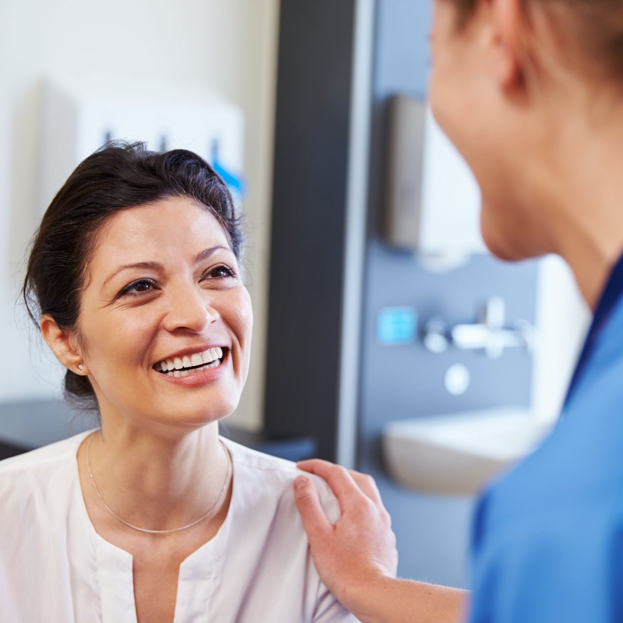 A smiling woman in a white shirt is comforted by a healthcare provider in a medical setting. The provider has a hand on the woman’s shoulder.