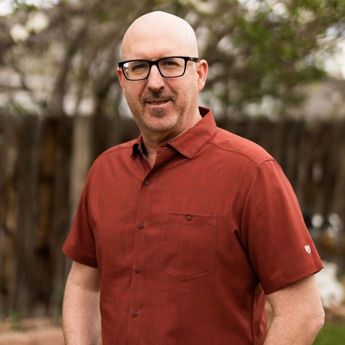 Man wearing glasses and a rust-colored shirt standing outdoors in front of trees
