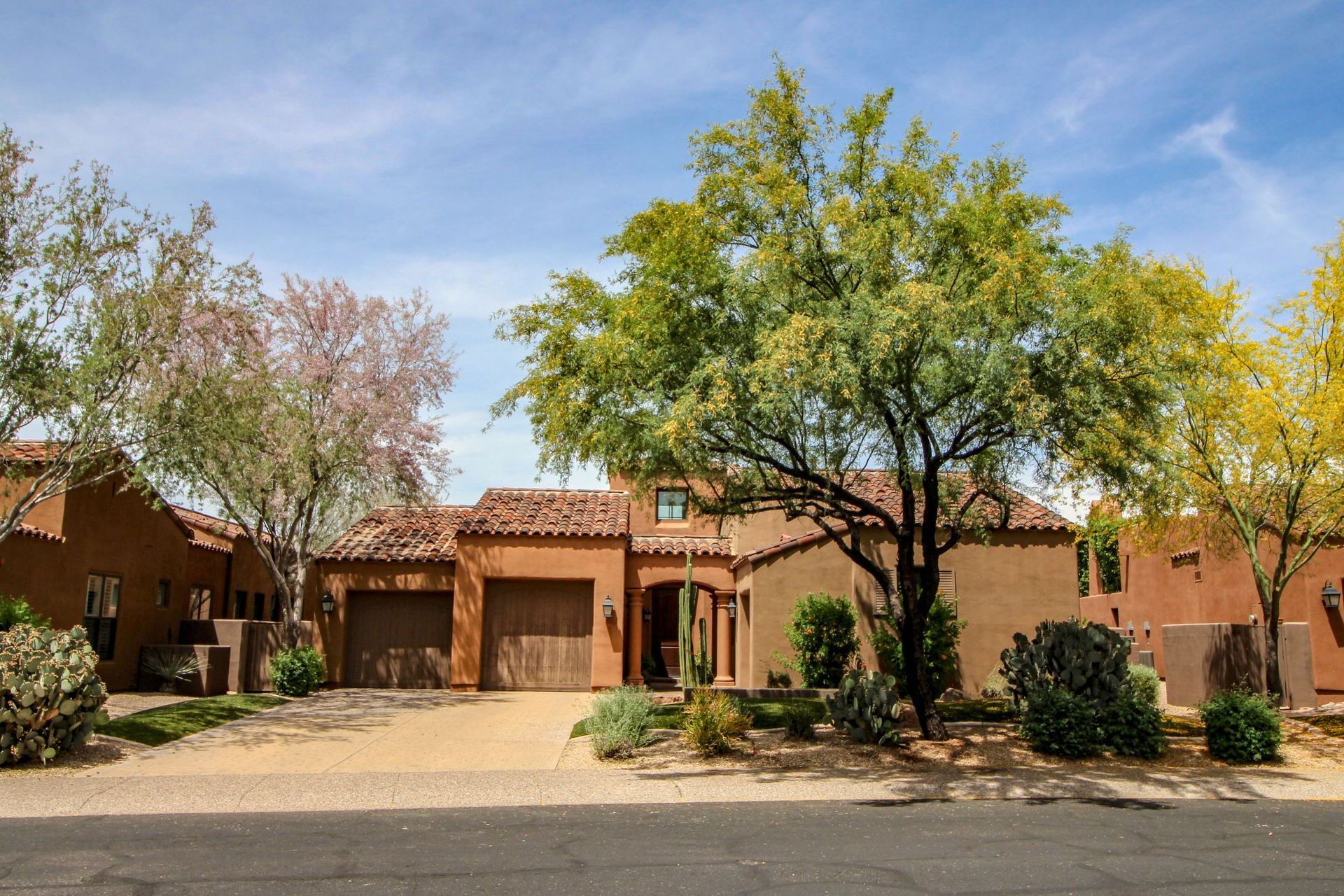 Suburban adobe-style house with a garage, desert landscaping, and trees under a blue sky
