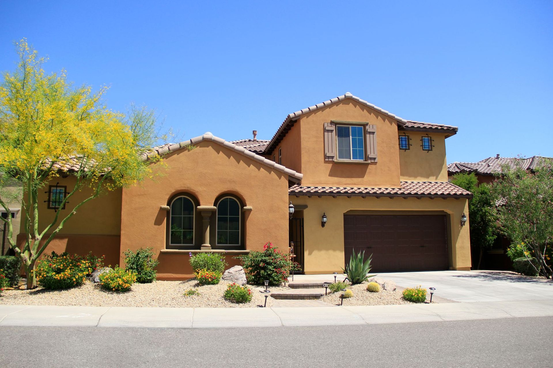 Two-story tan stucco house with a brown garage and desert landscaping under a clear sky